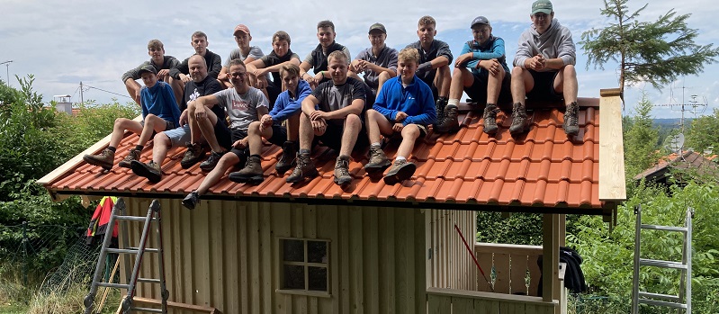 Schüler und Lehrkraft sitzen auf dem Dach einer Holzhütte