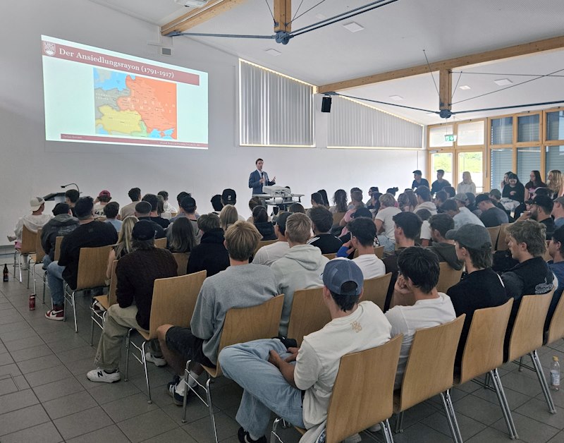 Jugendliche sitzen auf Stühlen und betrachten eine Leinwand, vorne steht ein Erwachsener am Pult.